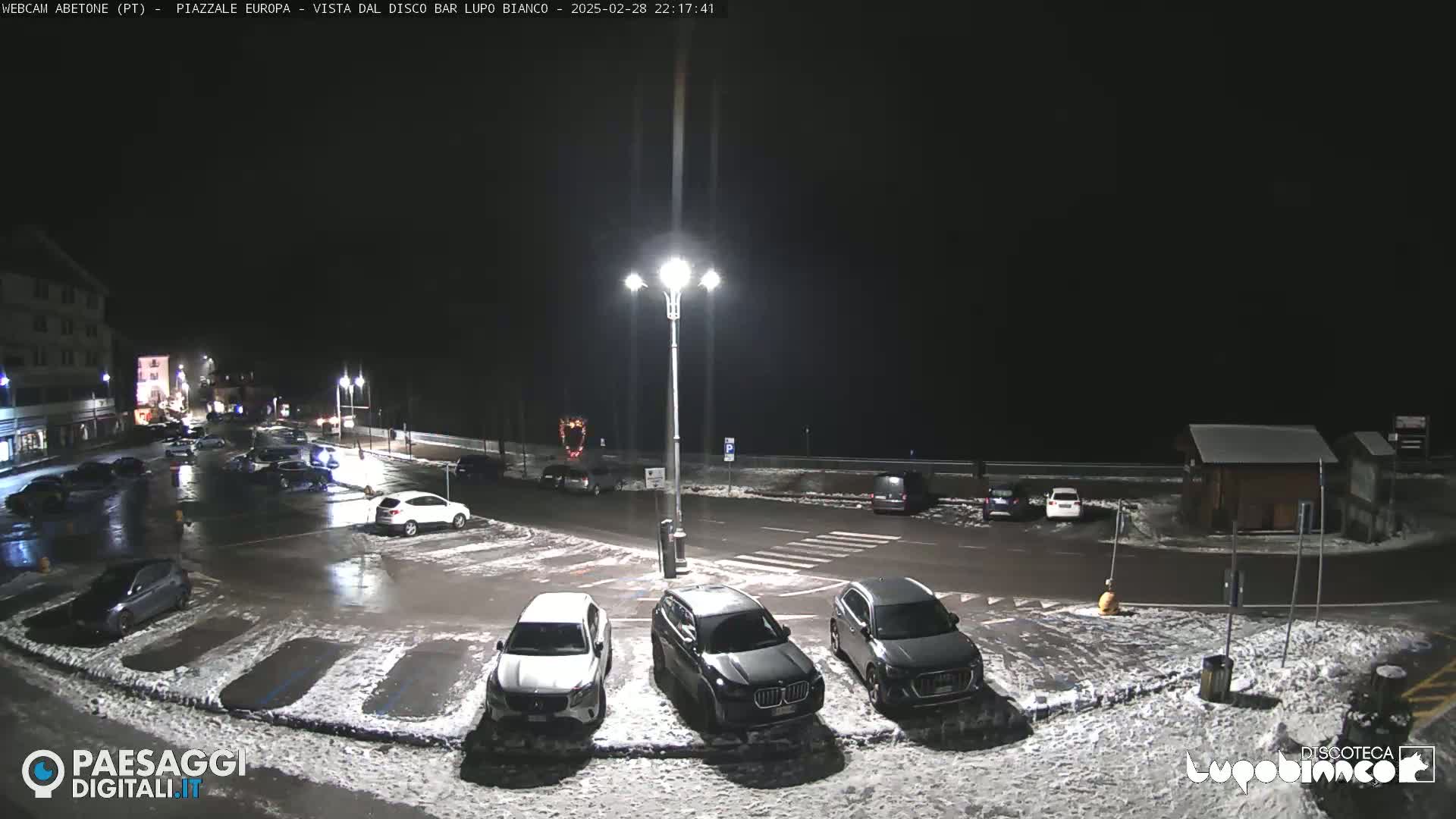 A nighttime view of a mostly empty parking lot with several cars, light snow on the ground, and streetlights illuminating the scene.