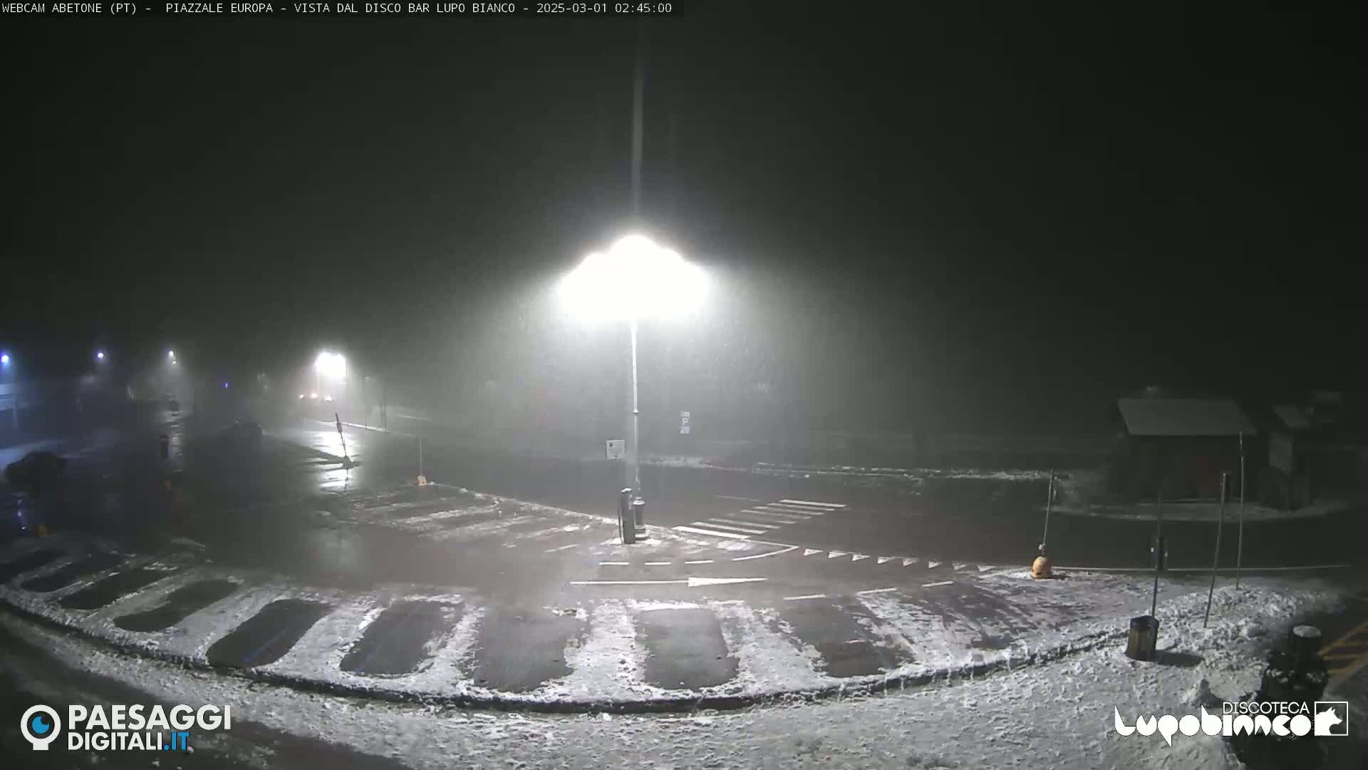 A nighttime, snowy scene shows a mostly empty parking lot with a street intersection, illuminated by bright streetlights under a foggy sky.