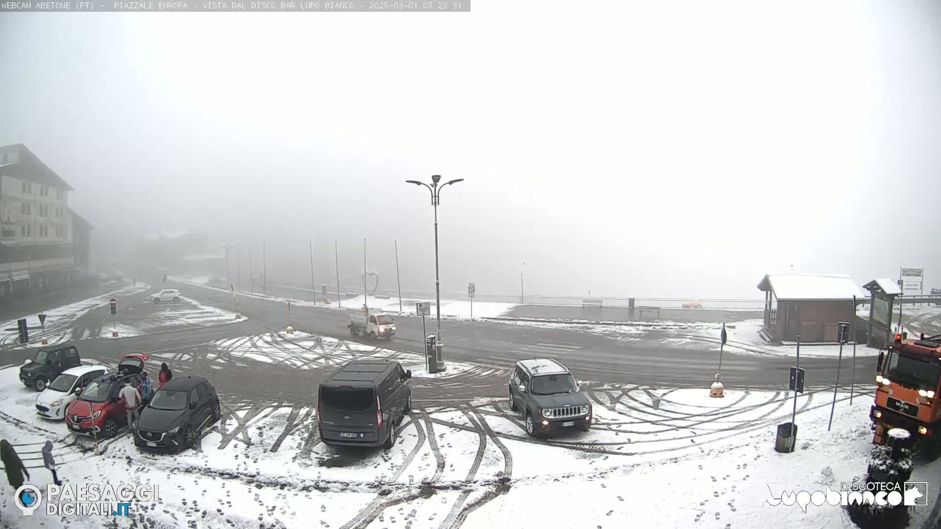 A snow-covered parking lot with several cars and a building in the background under foggy conditions.