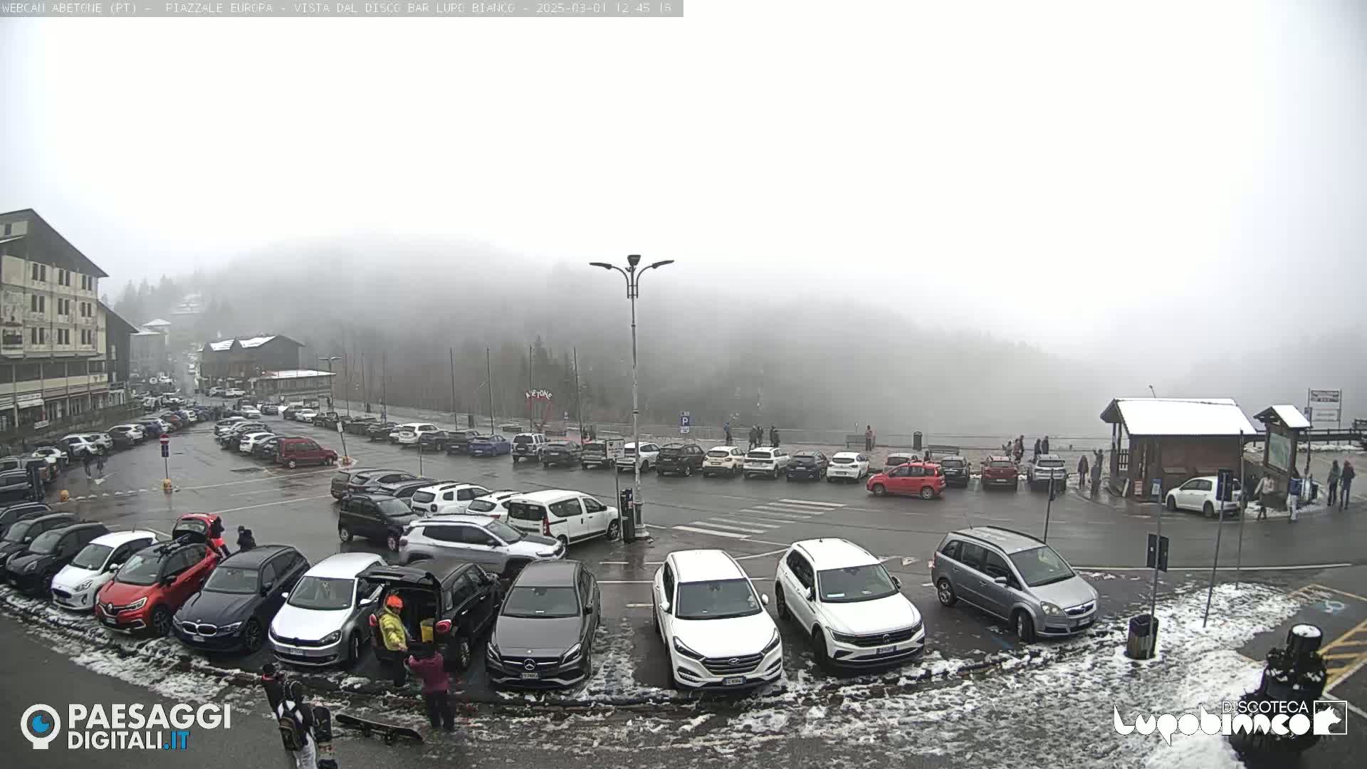 A foggy parking lot filled with cars, with a few people walking around and snow patches on the ground.