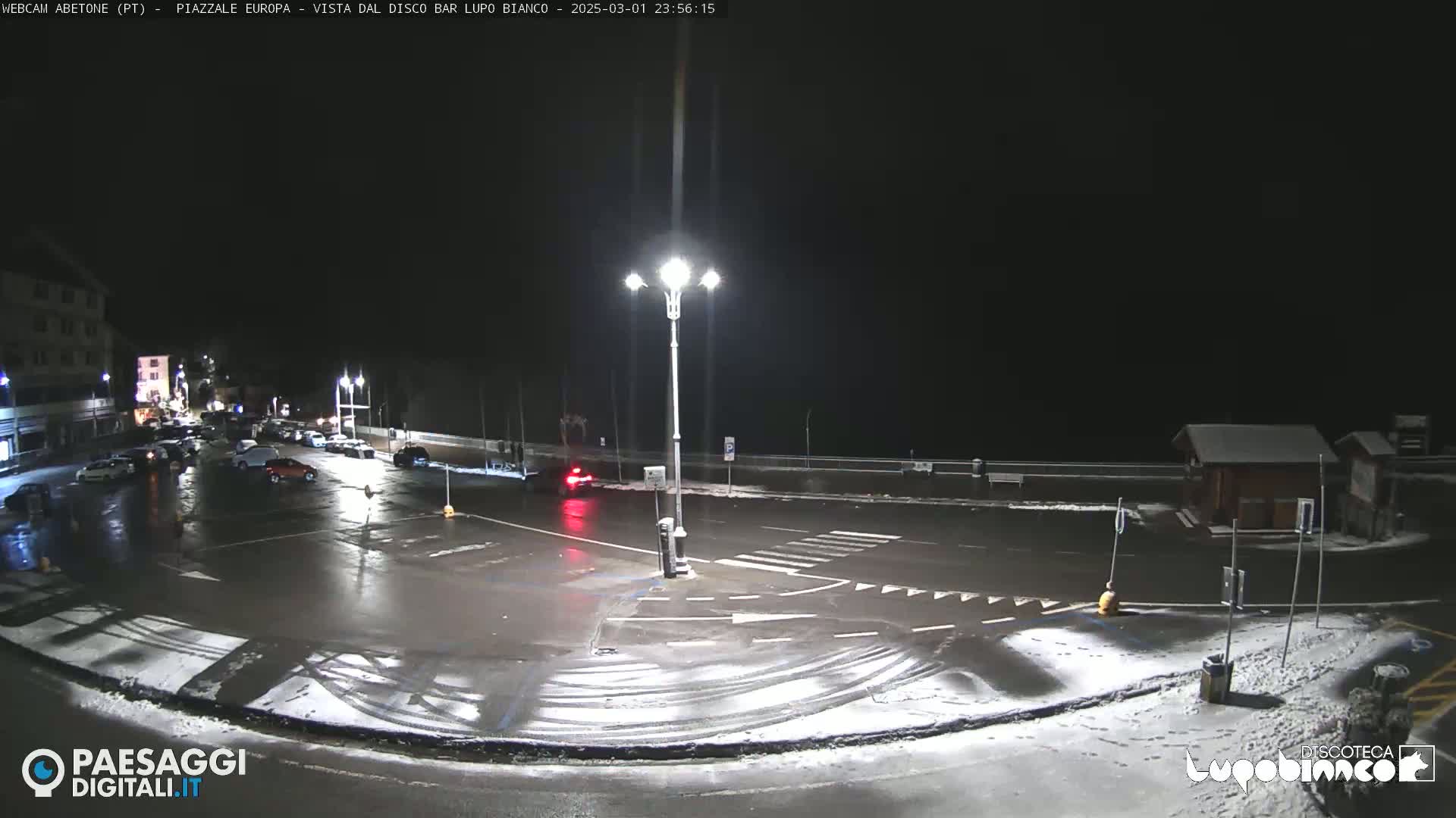 A nighttime, snowy scene shows a mostly empty parking lot and roadway illuminated by streetlights.