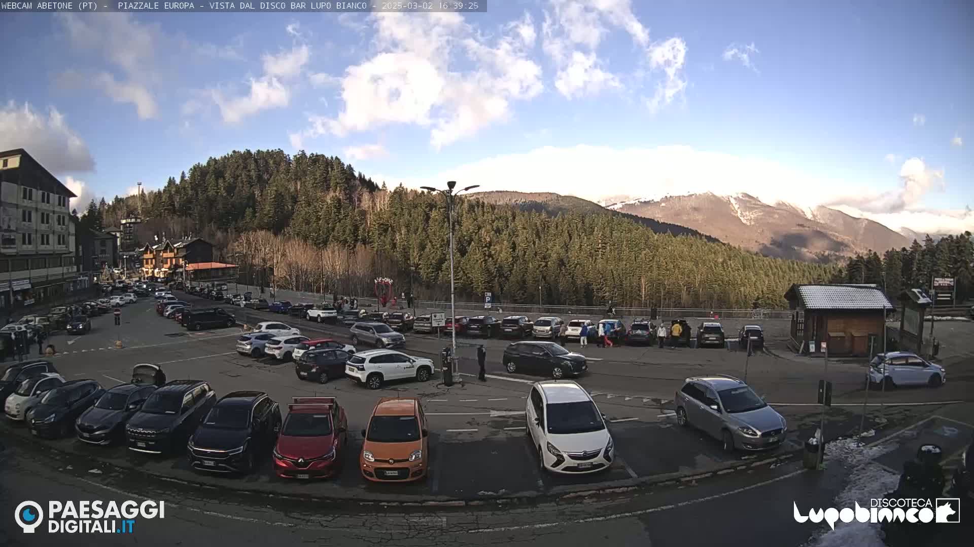 A mostly sunny day reveals a full parking lot surrounded by evergreen-covered mountains, with a few snow-dusted peaks visible in the distance.