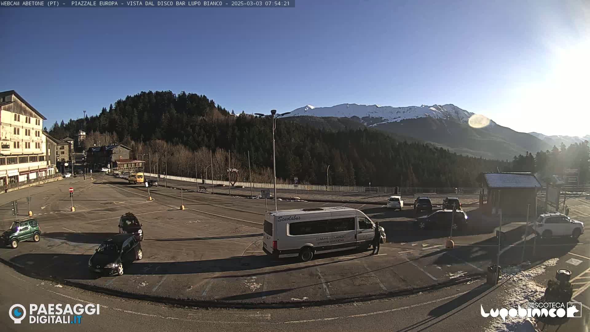 A mostly empty parking lot with several cars and a van, situated in front of buildings and trees, with snow-capped mountains in the background under a clear, sunny sky.