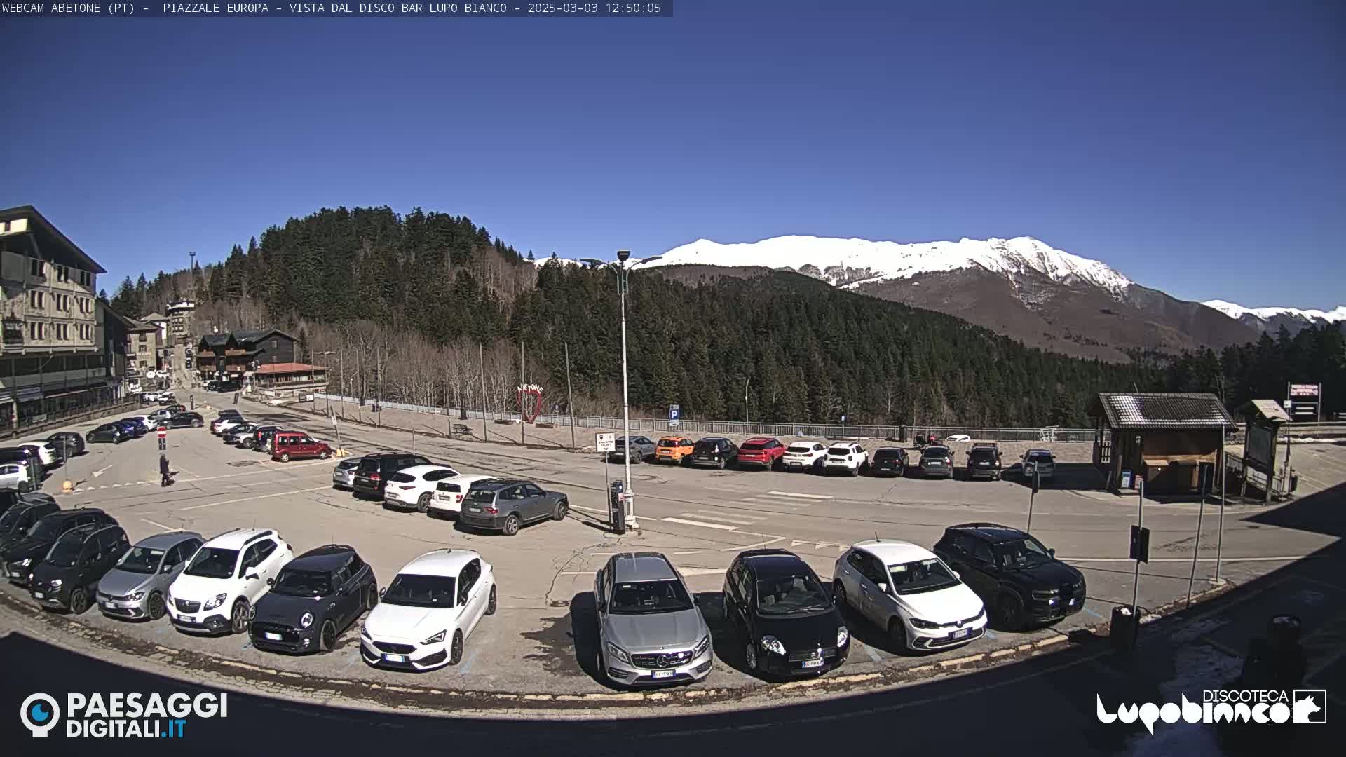 A mostly full parking lot sits before a mountain range with snow-capped peaks under a clear blue sky.