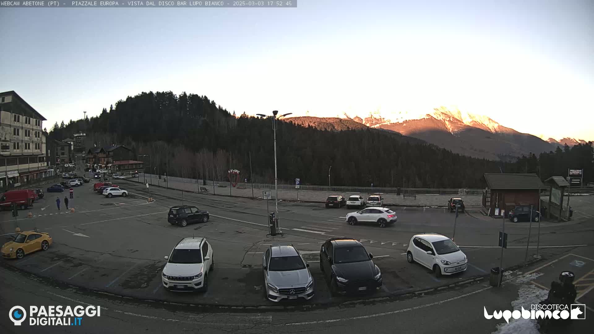 A mostly empty parking lot with several cars, situated in front of a building and surrounded by a mountainous landscape bathed in the warm light of a setting sun.