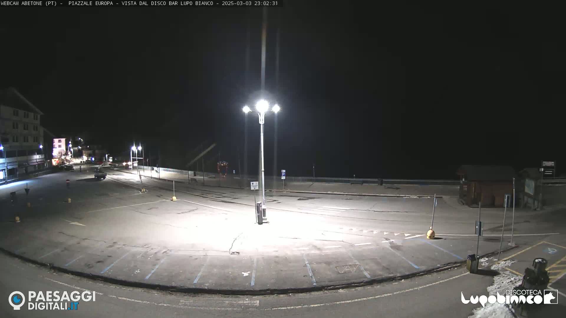 An empty, asphalt parking lot is illuminated at night by a tall lamppost, with a building visible in the background.