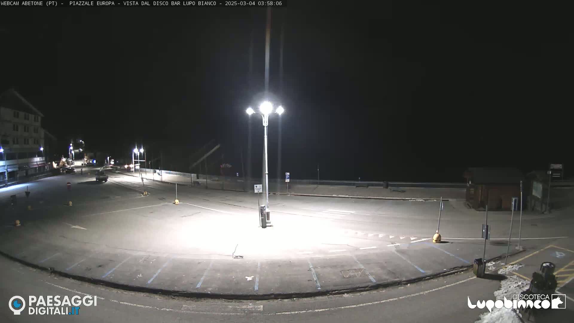 A mostly empty, brightly lit parking lot at night is shown, with a building and some vehicles visible in the distance.