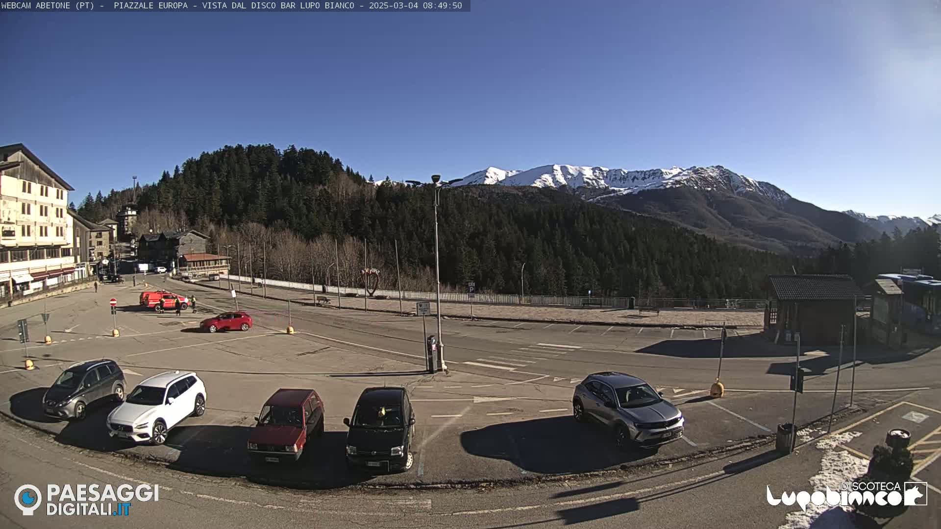 A mostly empty parking lot with several cars sits in front of a building and a backdrop of snow-capped mountains under a clear blue sky.