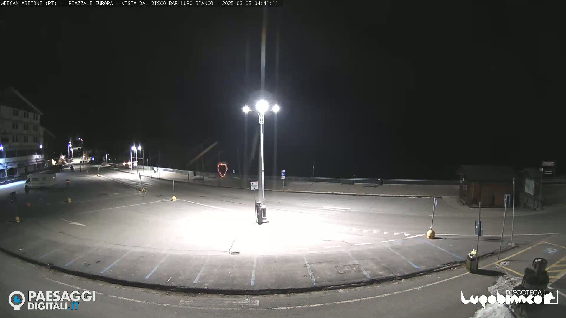 An empty, asphalt parking lot is illuminated by bright streetlights at night.