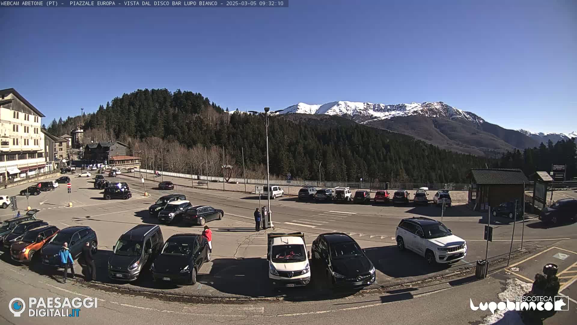A parking lot filled with cars is situated in front of a building, with snow-capped mountains and a forest visible in the background under a clear blue sky.