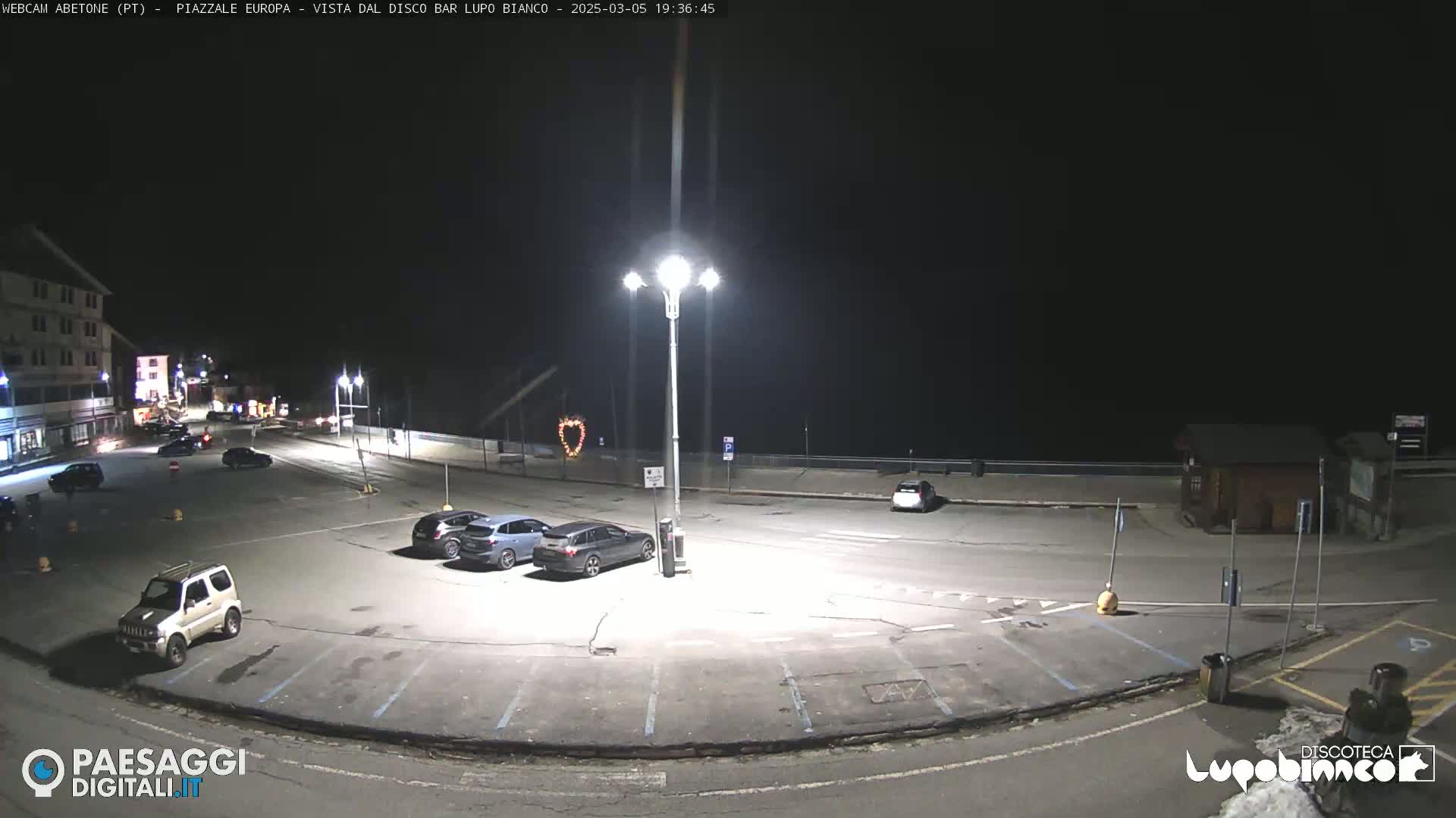 A nighttime view of an empty parking lot with a few cars, illuminated by streetlights, next to a building.