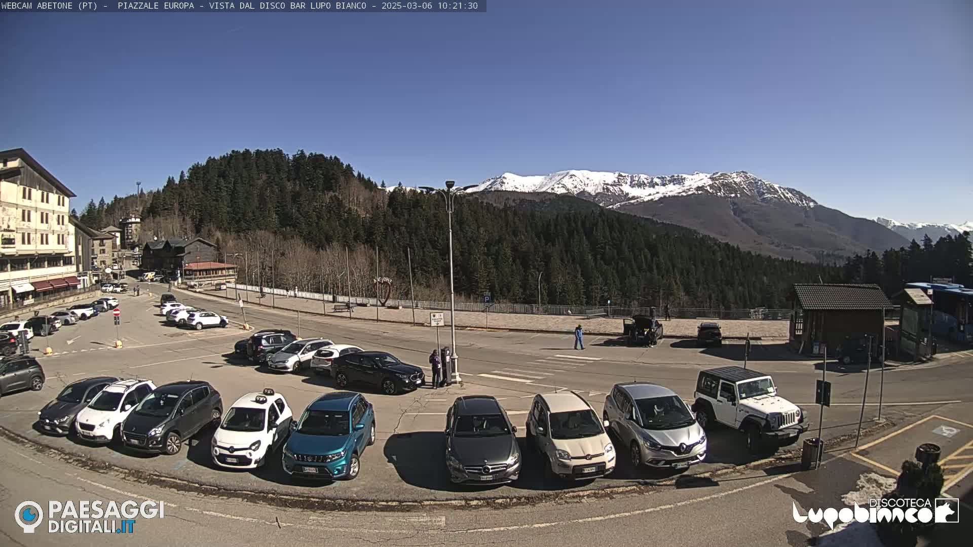 A parking lot filled with cars sits below a mountain range capped with snow under a clear blue sky.