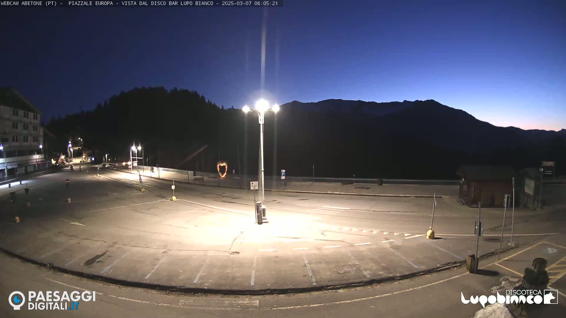 An empty, dark parking lot at twilight is illuminated by several streetlights, with a mountain range silhouetted against a dusky blue sky in the background.