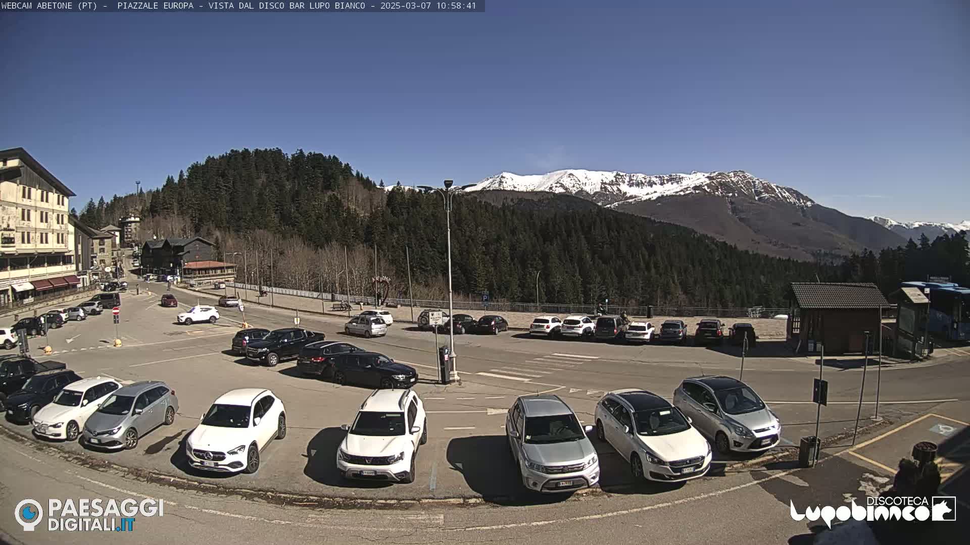 A parking lot with numerous cars is situated in front of a building and a mountain range with snow-capped peaks under a clear blue sky.