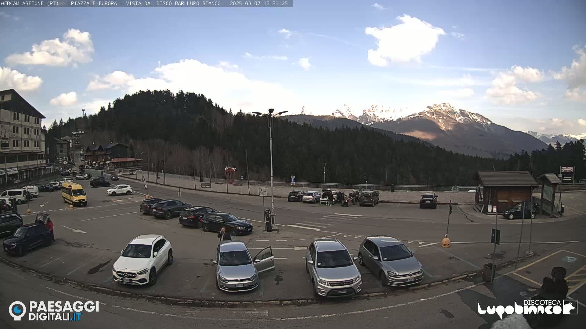 A parking lot with several cars is situated in front of a mountain range under a partly cloudy sky.