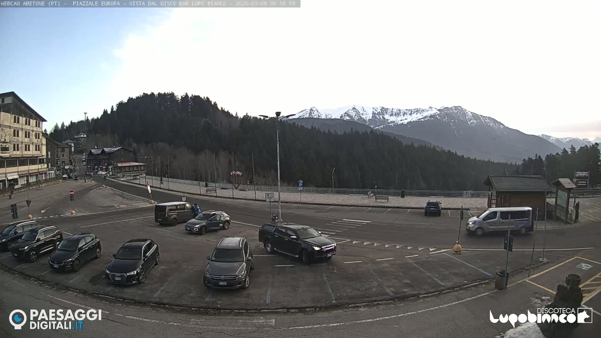 A mostly empty parking lot with several cars and a van is situated in front of a mountain range capped with snow under a mostly clear sky.