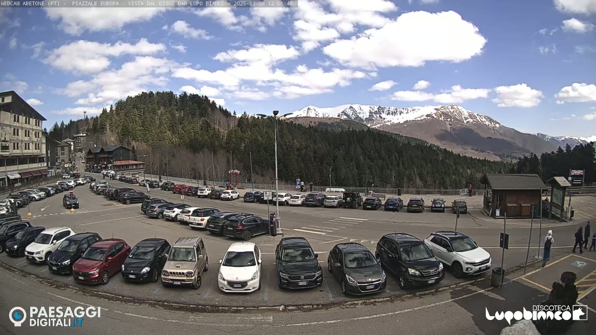 A sunny day with partly cloudy skies shows a large parking lot full of cars in front of a mountain range with snow-capped peaks and a dark evergreen forest.