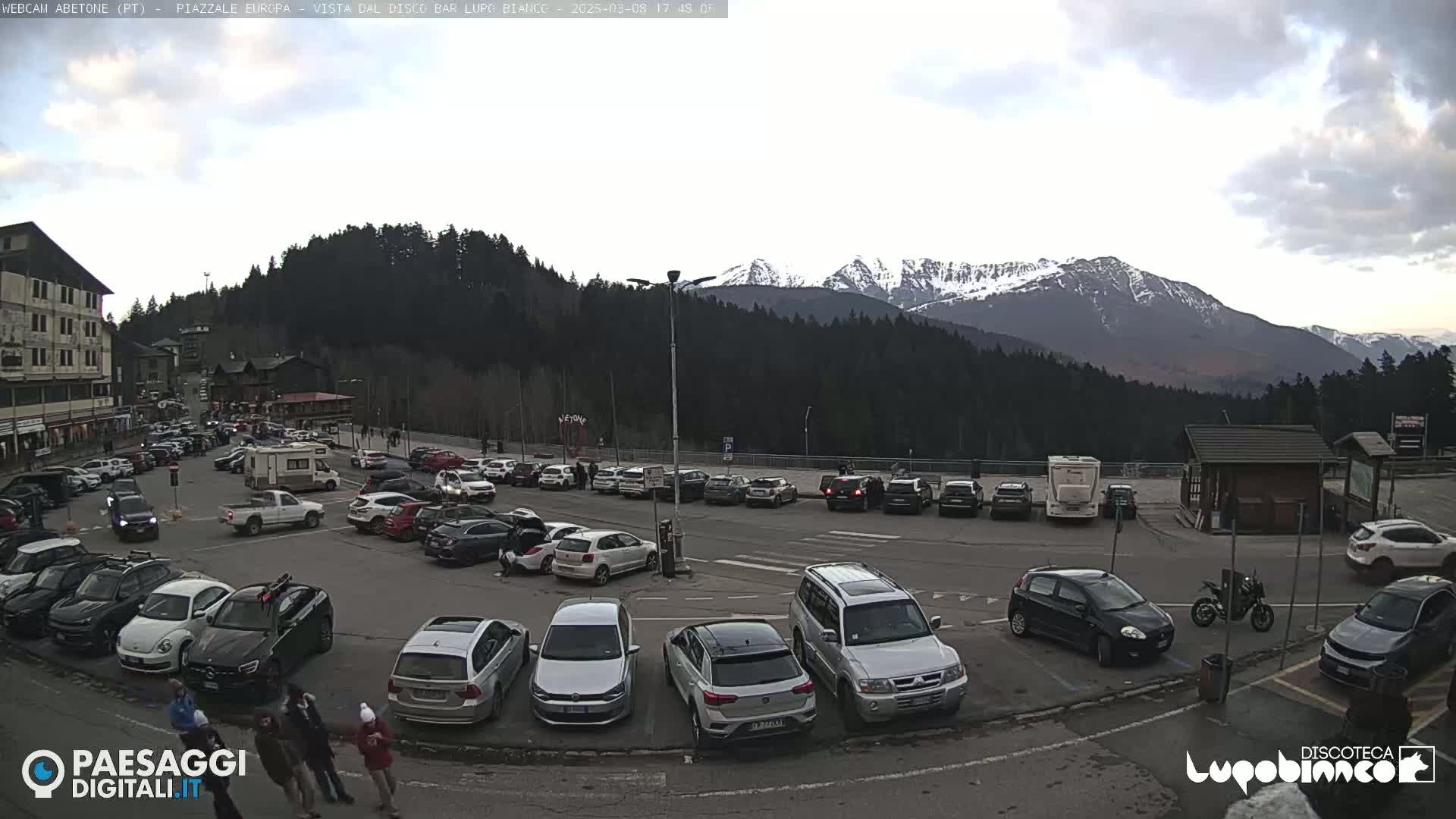 A parking lot filled with cars is situated below a mountain range with snow-capped peaks under a cloudy sky.