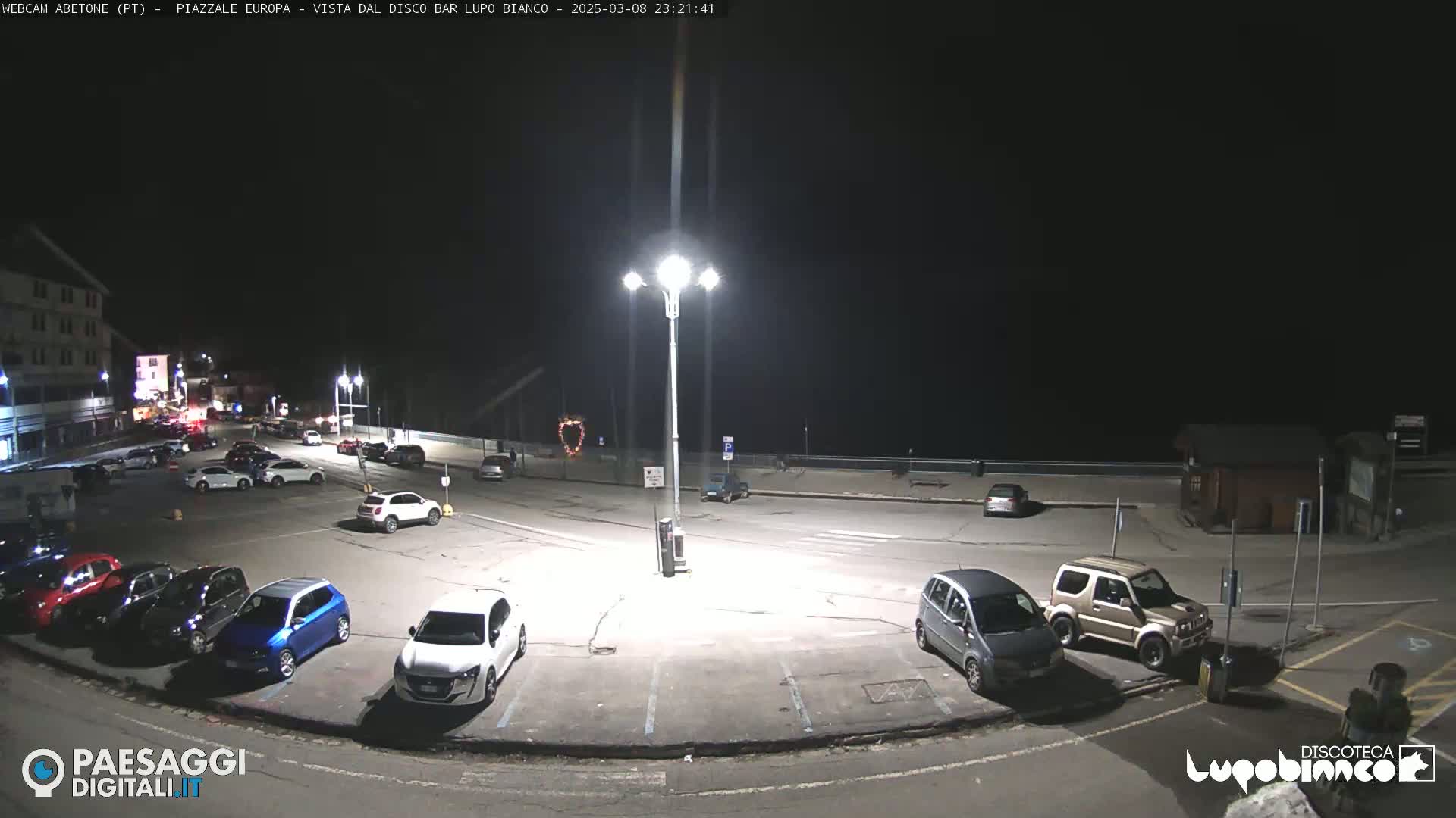 A nighttime view of a mostly empty parking lot with several cars parked, illuminated by a tall streetlight, next to a dark body of water.
