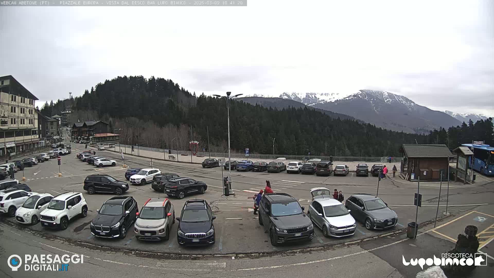 A mostly cloudy day in a mountain town parking lot filled with cars, with snow-capped mountains visible in the background.