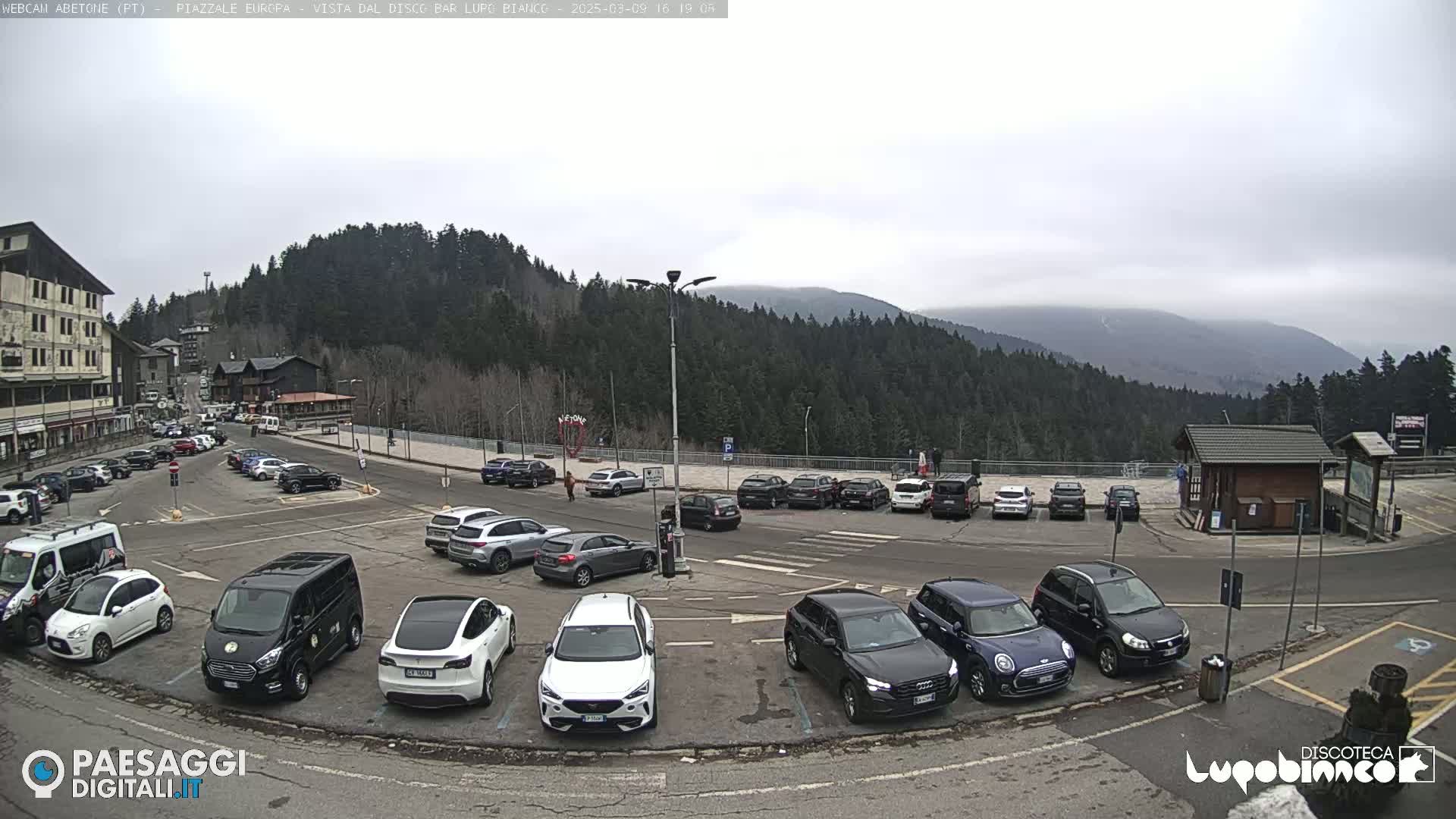 A parking lot filled with cars is situated in front of a mountain range under an overcast sky.