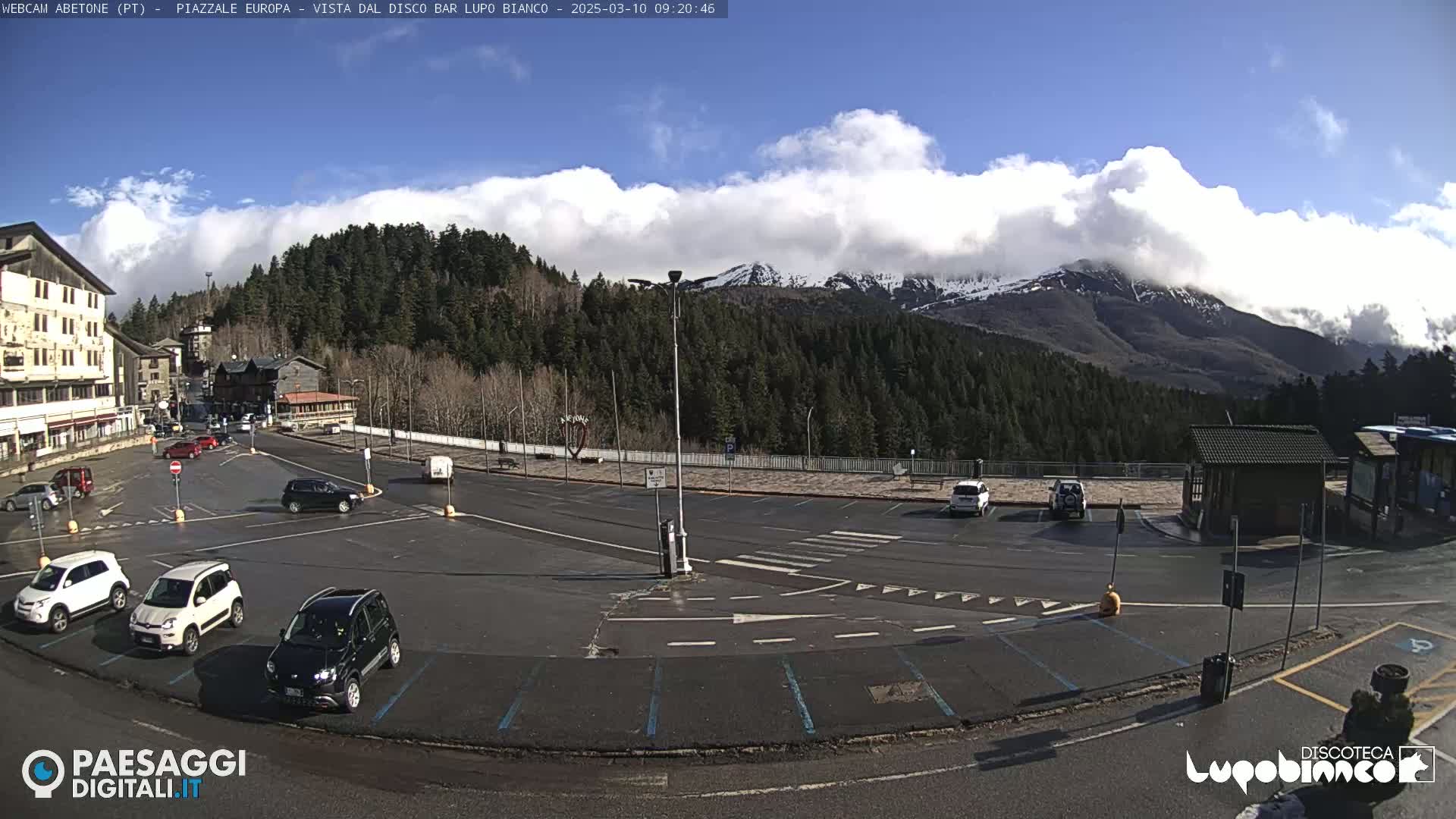 A mostly sunny day with partly cloudy skies shows a parking lot with several cars, a few buildings, and a mountainous backdrop with snow-capped peaks in the distance.