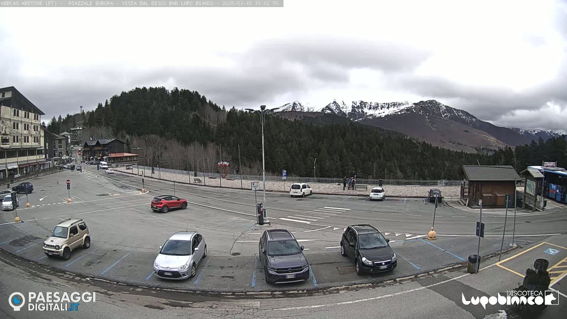 A parking lot with several cars is situated in front of a building, overlooking a mountain range with snow-capped peaks under a cloudy sky.