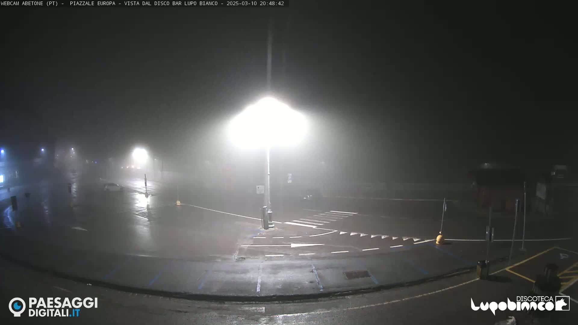 A nighttime, foggy view of a wet parking lot and road intersection illuminated by streetlights.