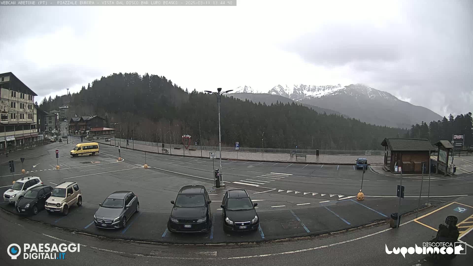 A mostly empty parking lot with several cars and a small yellow bus is situated in front of buildings and a snow-capped mountain range under an overcast sky.