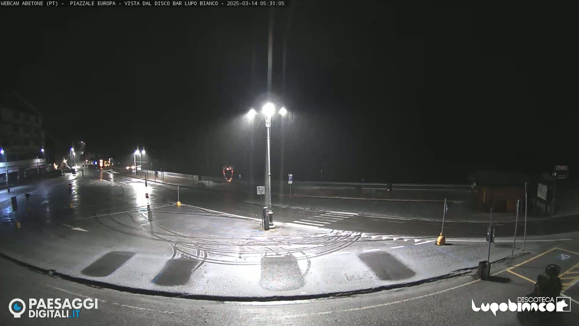 A nighttime, rain-slicked roadway intersection with minimal traffic under streetlights, showing some light snow accumulation.