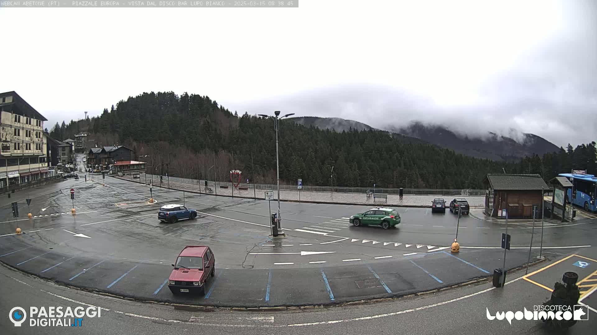 A wet parking lot with several cars, including a police car, is situated in front of a building and a wooded mountain range under an overcast sky.