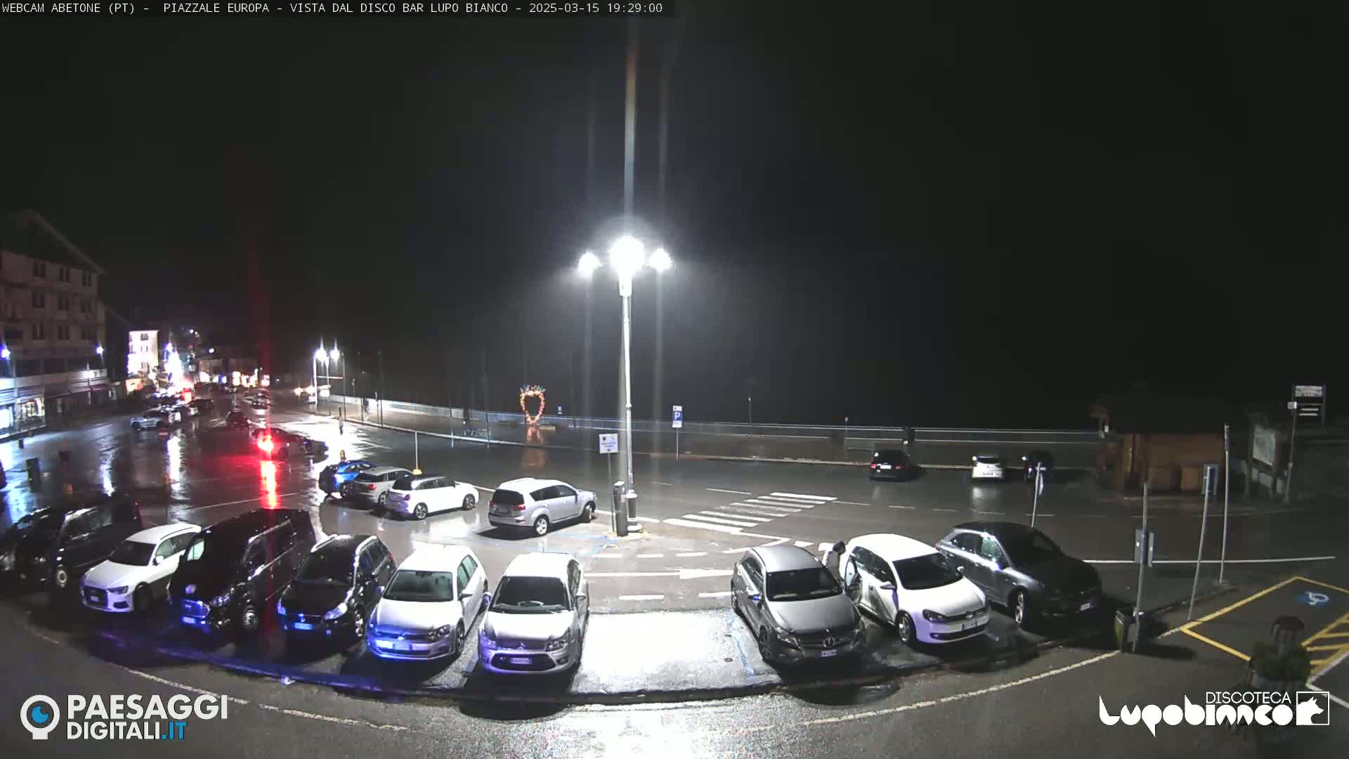 A nighttime, rain-soaked parking lot with several cars parked, illuminated by streetlights, near a waterfront.