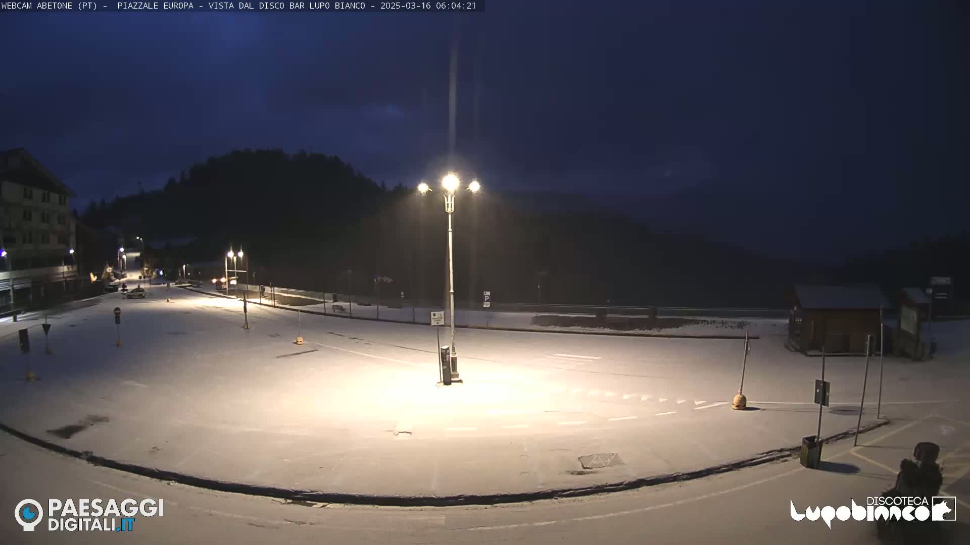 A snow-covered empty plaza is illuminated by streetlights at night, with dark mountains visible in the background.