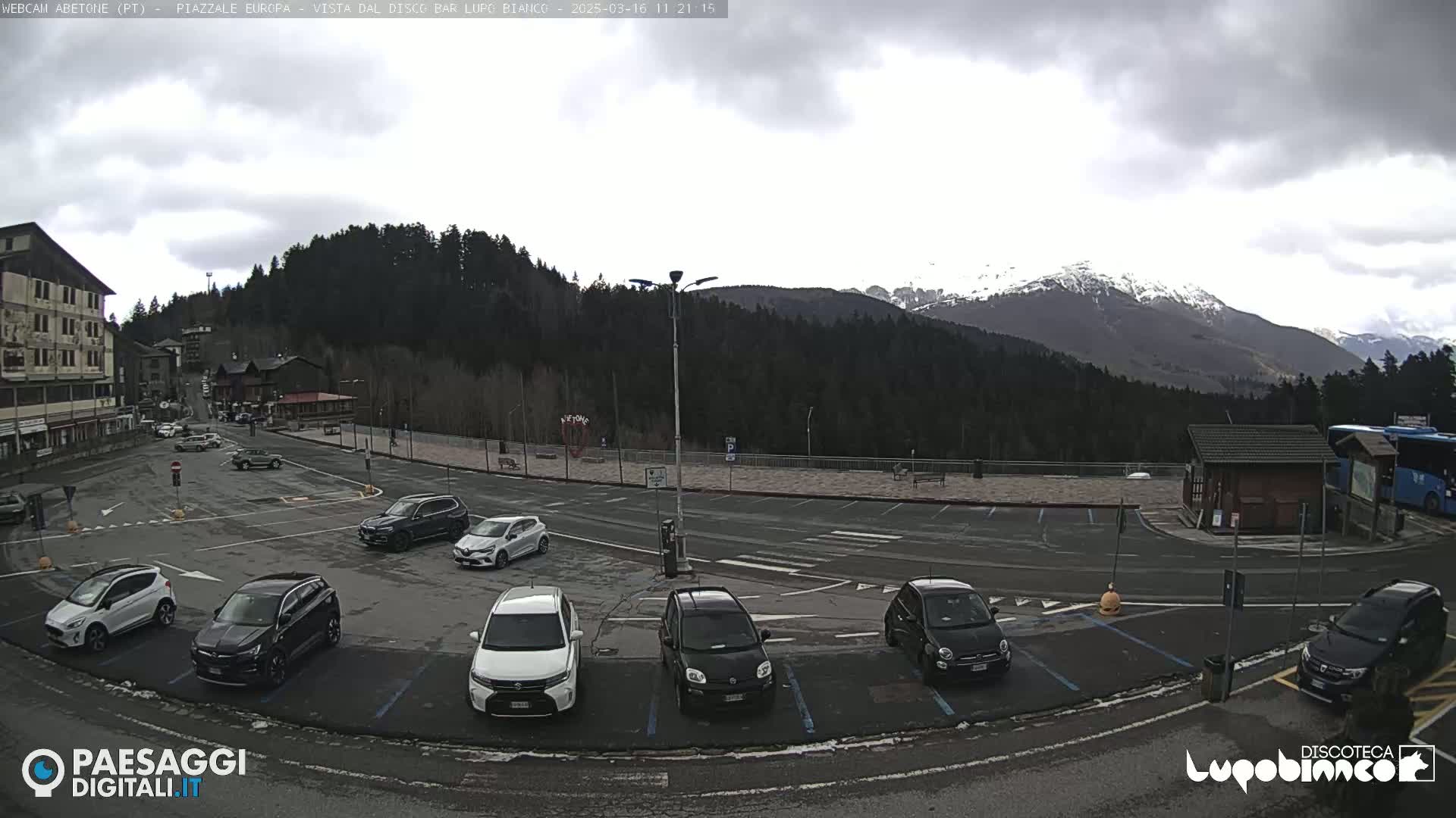 A mostly cloudy day shows a parking lot with several cars, a building, and a mountainous background with snow-capped peaks.
