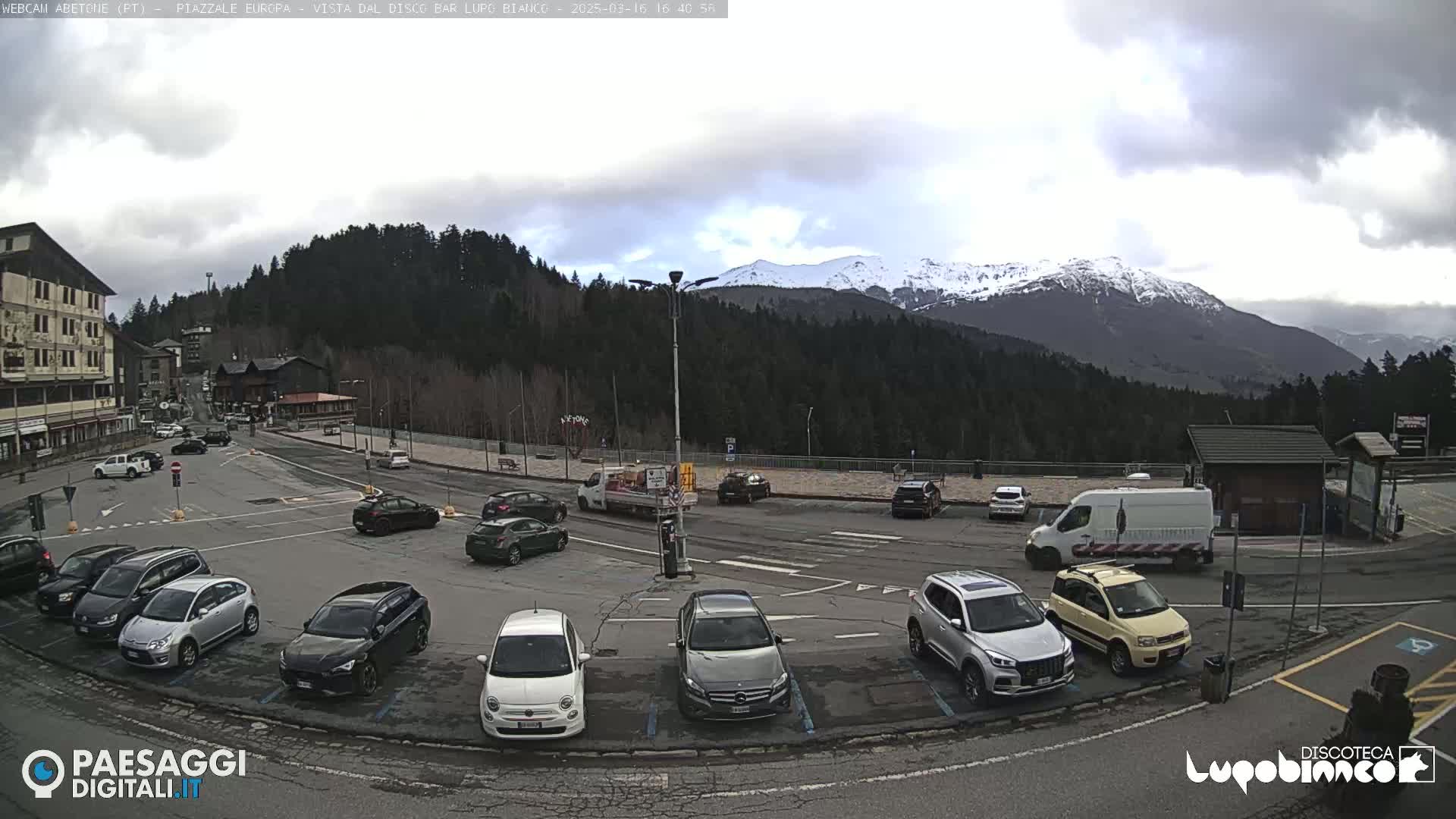 A parking lot with numerous cars is situated near a roadway, backed by a wooded mountain range with snow-capped peaks under a cloudy sky.