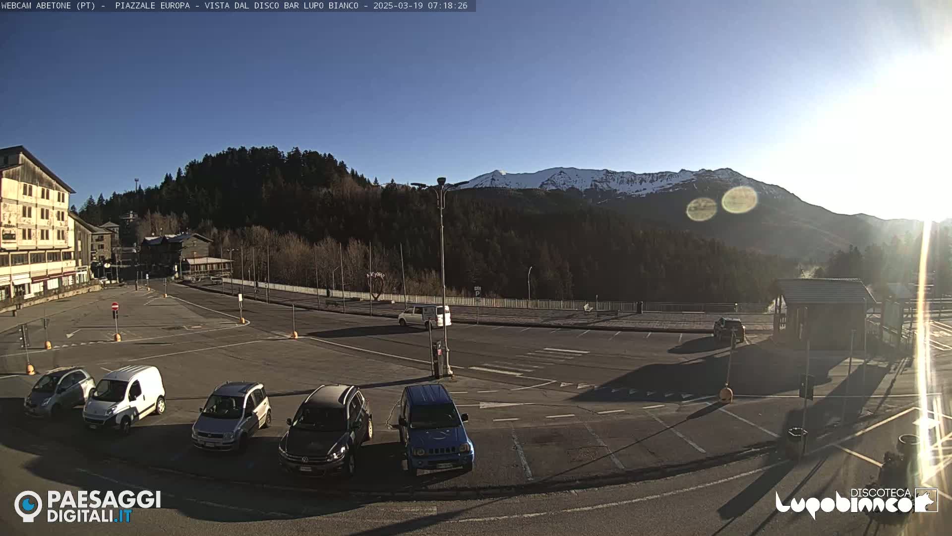 A mostly empty parking lot with several cars, situated in front of buildings and a wooded hillside with snow-capped mountains in the background, under a clear sunny sky.