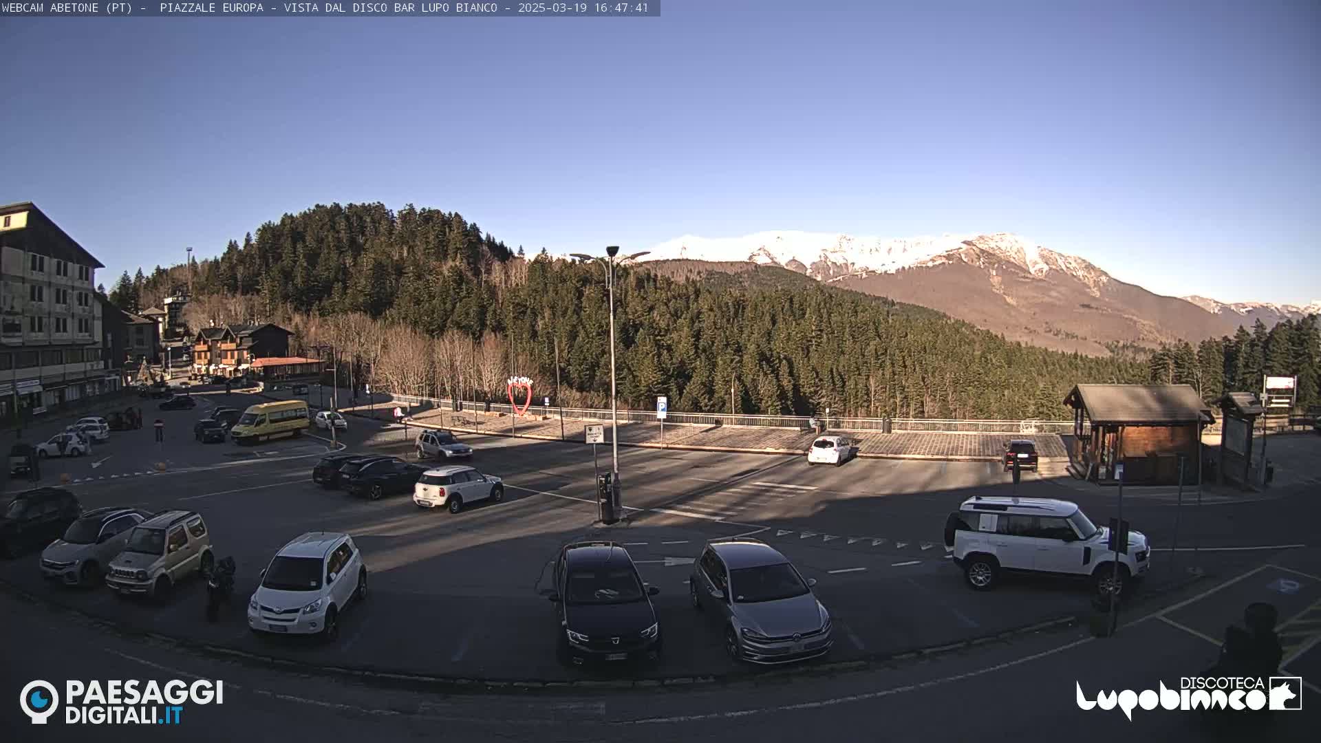 A sunny, snow-capped mountain range overlooks a mostly empty parking lot with several cars and a bus, situated in front of a building and a wooded area.
