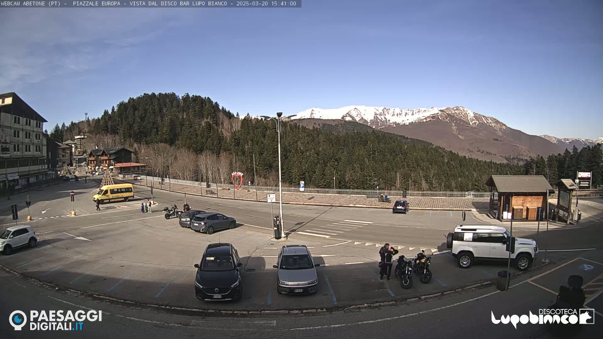 A sunny day in a mountain town parking lot with several cars, a school bus, and motorcycles parked, along with a few pedestrians; snow-capped mountains are visible in the background.
