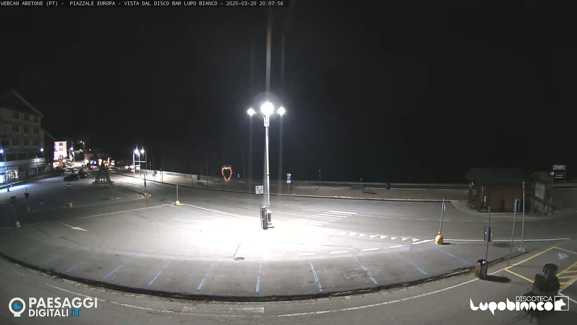 An empty, dark parking lot and roadway are illuminated by bright streetlights at night.