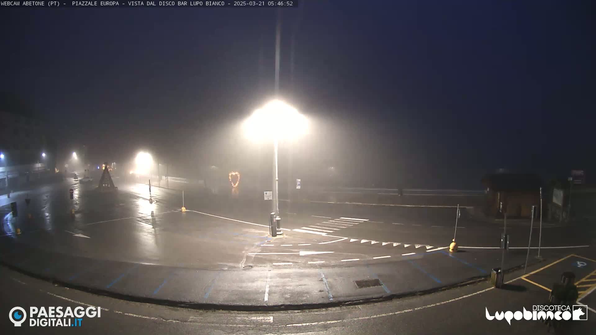 A foggy, rain-slicked empty parking lot is illuminated by streetlights at night.