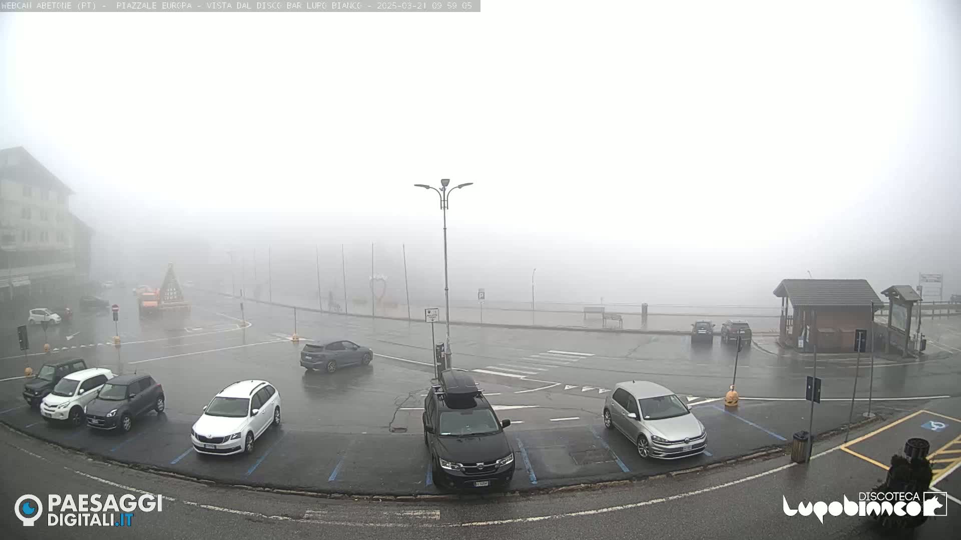 A foggy, wet parking lot with several cars parked, situated in front of a building and a small structure.
