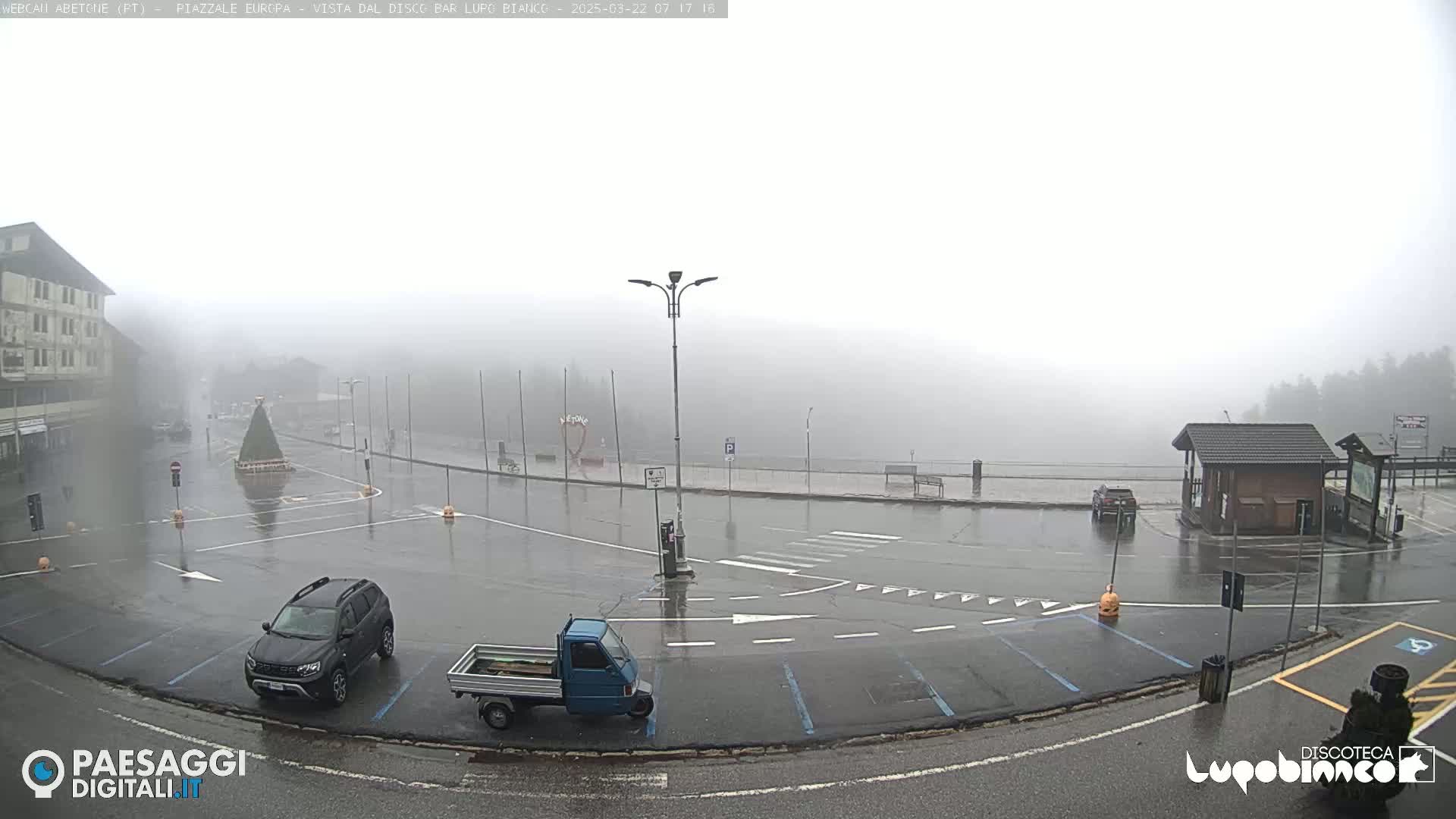 A mostly empty, wet parking lot is shrouded in a thick fog, with a few vehicles present and a building in the background.