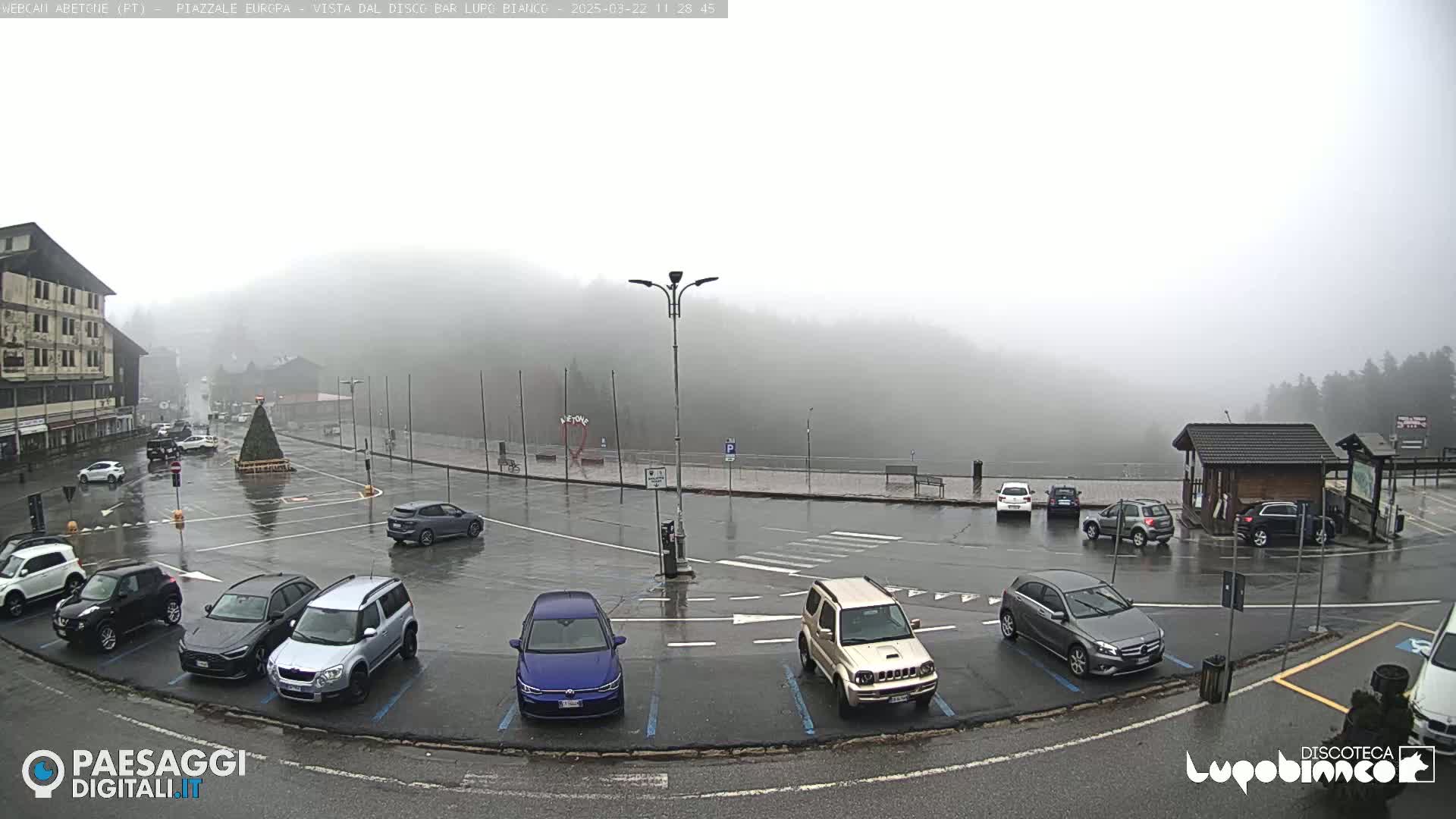 A foggy, rainy parking lot with several cars parked, surrounded by buildings and a forested hill in the background.