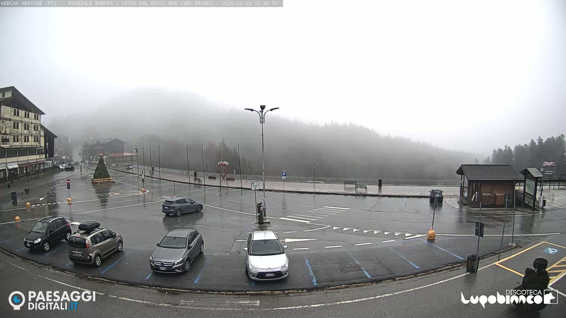 A wet, foggy parking lot with several cars is surrounded by buildings and a forested hill.