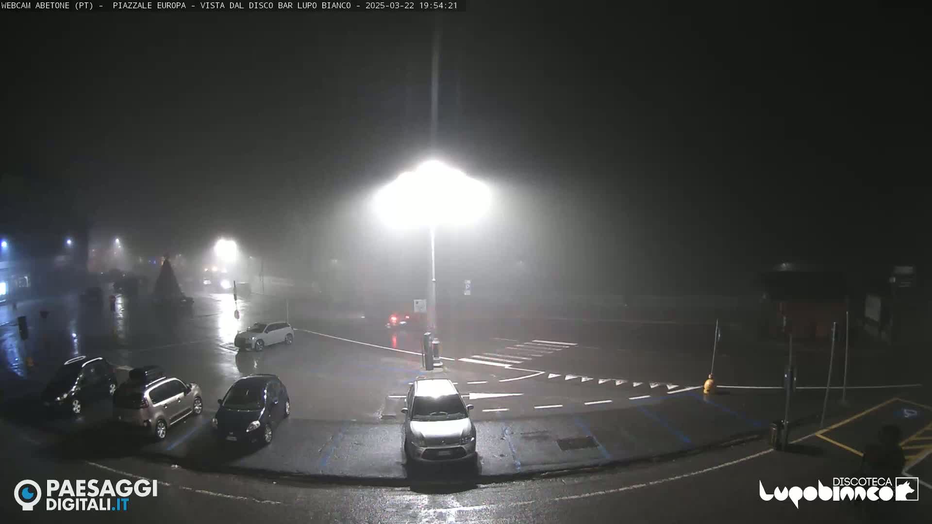 A nighttime, rainy scene shows a mostly empty parking lot with several cars parked and a few driving, illuminated by bright streetlights in hazy conditions.