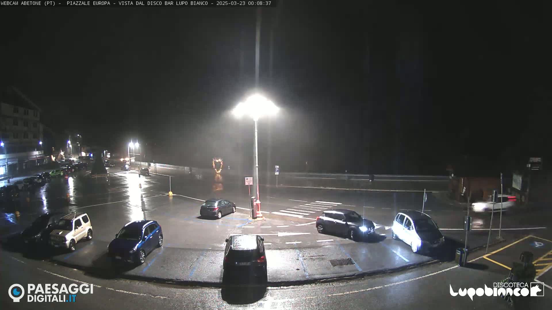 A rain-slicked nighttime parking lot with several cars parked and a few driving by, illuminated by bright streetlights.