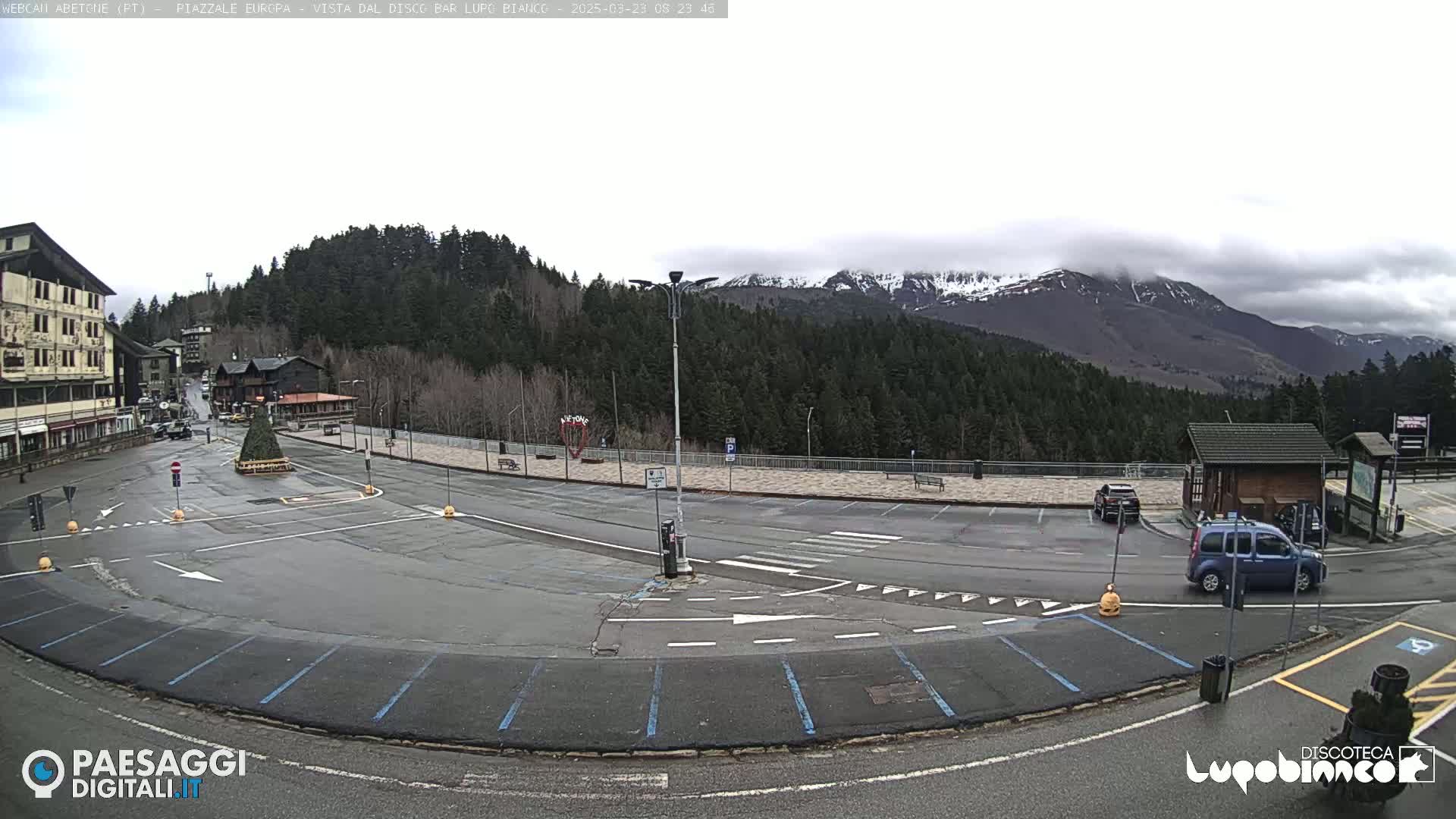 An overcast day shows a mostly empty parking lot and roundabout near a building and snow-capped mountains in the background.