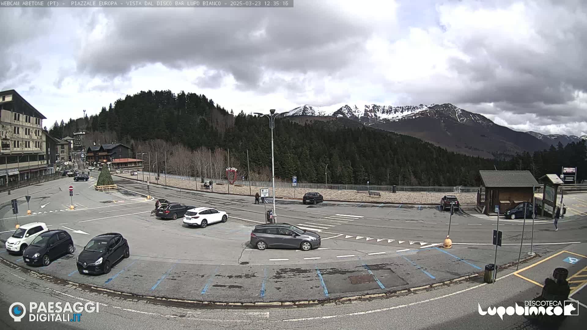 A mostly cloudy day shows a parking lot with several cars, a few buildings, and snow-capped mountains in the background.