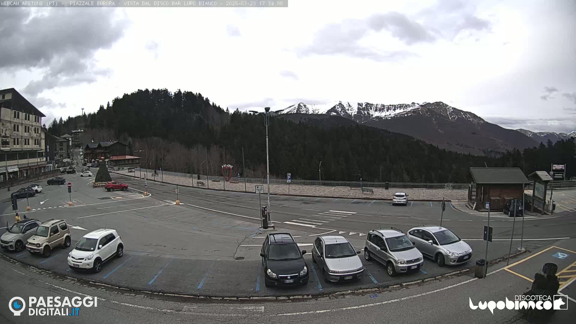 A mostly empty parking lot with several cars, situated in front of a building and a mountainous backdrop under an overcast sky.