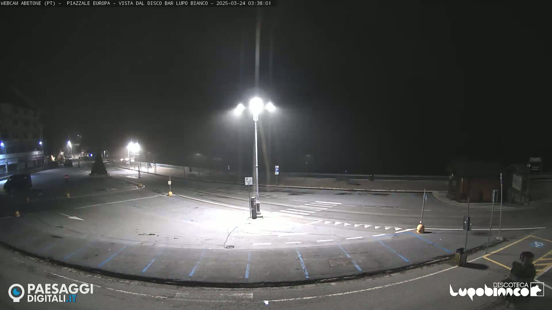 A mostly empty, paved plaza at night, dimly lit by streetlights, is shrouded in light fog.